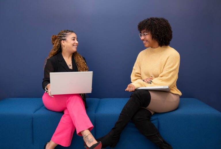 Two women sitting on a covered bench looking at each other and holding laptops.